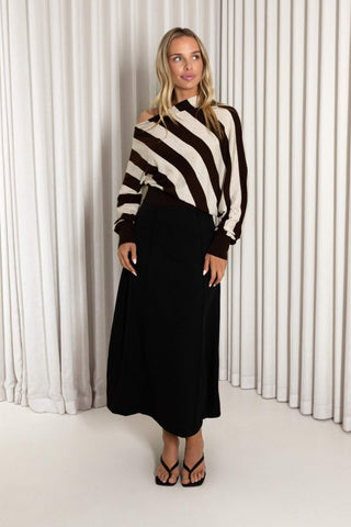 A woman modeling a brown and cream striped off-the-shoulder knit sweater paired with a black midi skirt and black strappy sandals, standing in front of a white curtain backdrop.