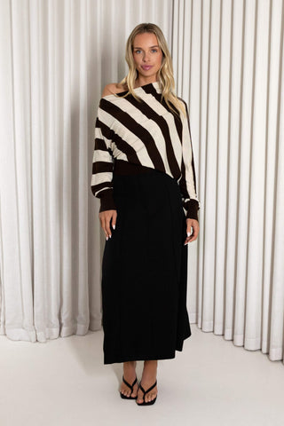 A woman modeling a long-sleeve knit top with bold diagonal stripes in brown and cream, paired with a long black skirt, standing in front of white pleated curtains.