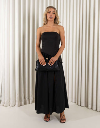 Woman wearing a black strapless dress with a slit, holding a black clutch against a white curtain background. Chris black dress with contrasting fabric and skirt split