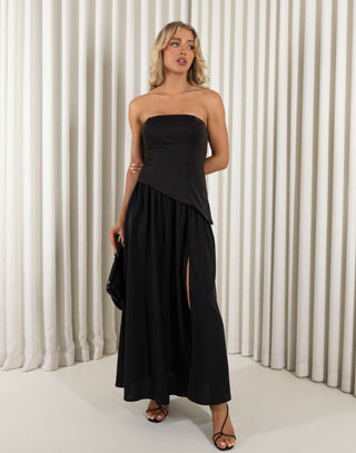 Woman wearing a black strapless dress with a slit, holding a black clutch against a white curtain background. Chris black dress with contrasting fabric and skirt split