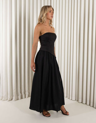 Woman wearing a black strapless dress with a slit, holding a black clutch against a white curtain background. Chris black dress with contrasting fabric and skirt split