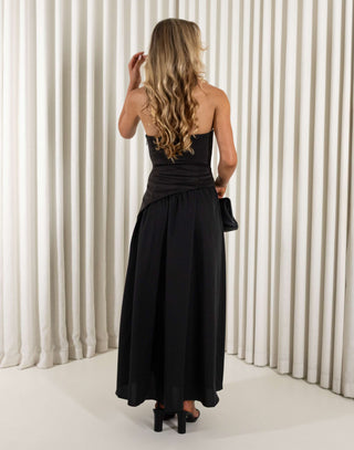 Woman wearing a black strapless dress with a slit, holding a black clutch against a white curtain background. Chris black dress with contrasting fabric and skirt split