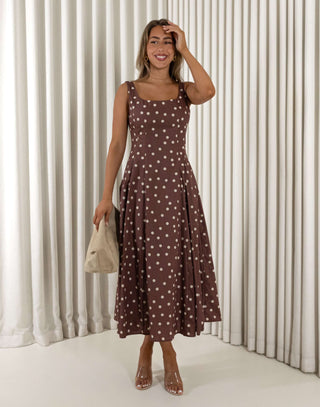 Woman wearing a brown polka dot dress holding a beige bag against a white curtain background Christine A-line midi dress in polka dots with squoval neckline
