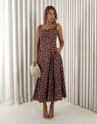 Woman wearing a brown polka dot dress holding a beige handbag against a white curtain background Christine A-line midi dress in polka dots with squoval neckline