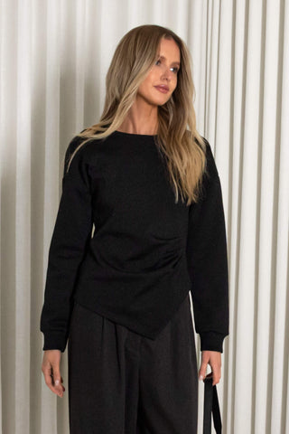A woman wearing a black long-sleeve knit top with an asymmetrical draped hem and black wide-leg trousers, standing in front of a beige pleated curtain backdrop.