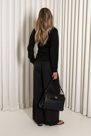 A woman with long blonde hair is shown from behind, wearing a black long-sleeve top and wide-leg black trousers, holding a black leather shoulder bag with a gold logo plate. She is standing in front of a white curtain backdrop.