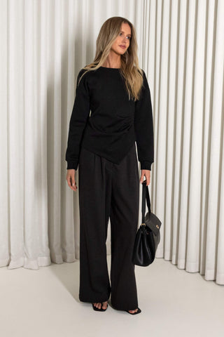 A woman modeling a black long-sleeve top and matching wide-leg pants, holding a black handbag, set against a background of white vertical drapes.