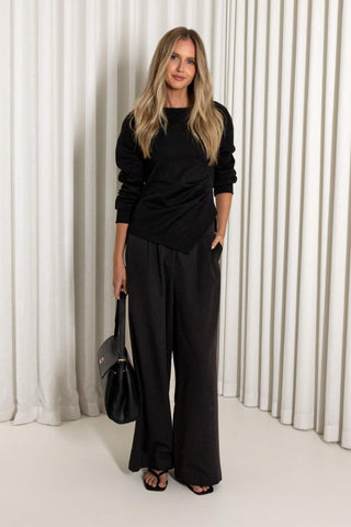 A woman stands in front of white pleated curtains wearing a black long-sleeve top and matching black wide-leg pants, holding a black handbag.