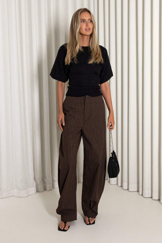 A woman stands in front of a white curtain backdrop wearing a black short-sleeve top and brown pinstriped wide-leg trousers, holding a small black handbag.