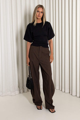 A woman stands in front of white pleated curtains wearing a black short-sleeve top and brown pinstriped wide-leg trousers, holding a small black handbag.
