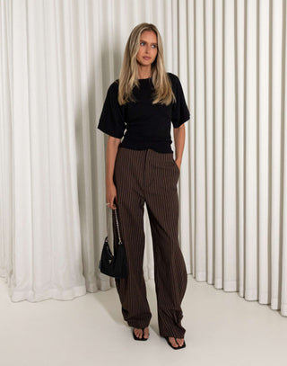 Woman wearing a black top and brown striped pants against a white curtain background