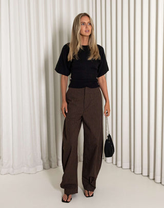 Woman wearing a black top and brown striped pants against a white curtain background