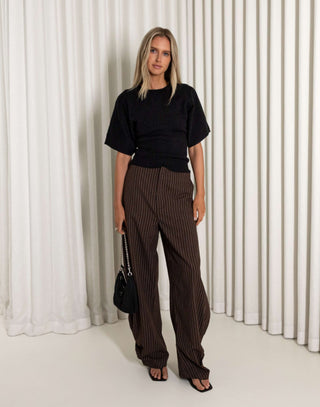 Woman wearing a black top and brown striped pants against a white curtain background