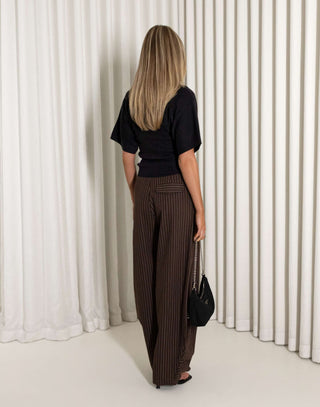 Woman wearing a black top and brown striped pants against a white curtain background