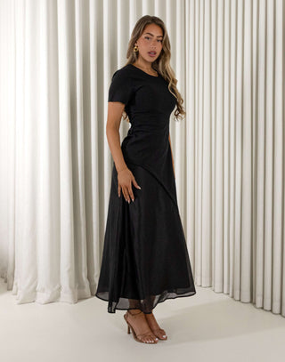 Woman wearing a black dress standing against a white curtain background Davina black contrast sheen maxi dress with short sleeves