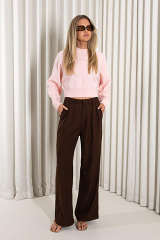 A woman modeling a light pink ribbed knit cropped sweater and dark brown wide-leg trousers, standing against a white curtain backdrop.