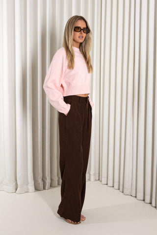A woman modeling a light pink ribbed knit cropped sweater and dark brown wide-leg trousers, standing against a white curtain backdrop.