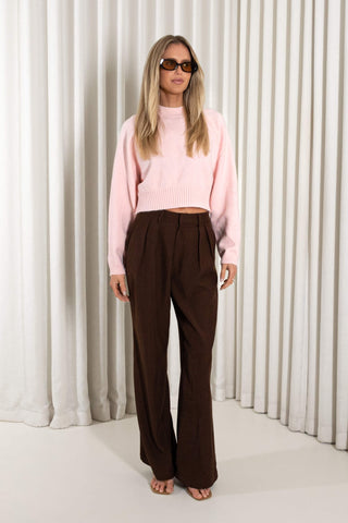 A woman modeling a light pink ribbed knit cropped sweater and dark brown wide-leg trousers, standing against a white curtain backdrop.