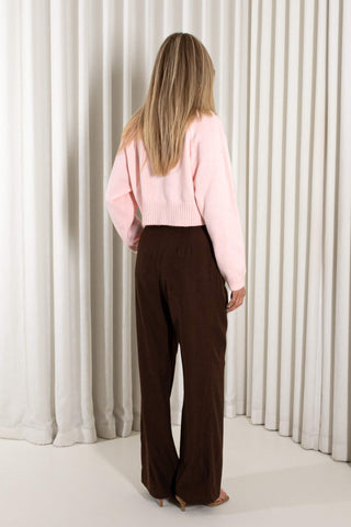 A woman modeling a light pink ribbed knit cropped sweater and dark brown wide-leg trousers, standing against a white curtain backdrop.