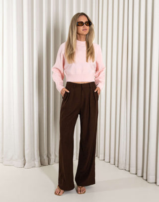 Woman wearing a pink sweater and brown pants against a white curtain background