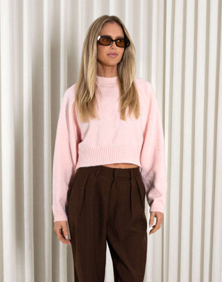 Woman wearing a pink sweater and brown pants against a white curtain background