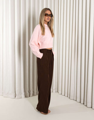 Woman wearing a pink sweater and brown pants against a white curtain background
