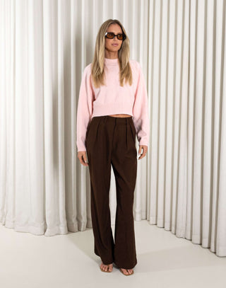 Woman wearing a pink sweater and brown pants against a white curtain background