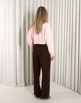 Woman wearing a pink sweater and brown pants against a white curtain background