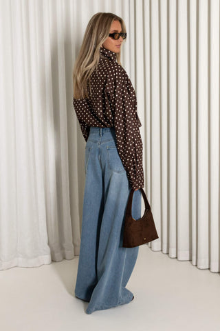 A woman stands in profile wearing a dark brown long-sleeve blouse with white polka dots, high-waisted wide-leg blue jeans, and holding a brown leather handbag, set against a background of white vertical drapes.