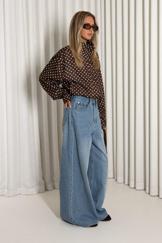 A woman modeling a brown long-sleeve blouse with white polka dots and high-waisted, wide-leg light blue denim jeans, standing in front of white pleated curtains.