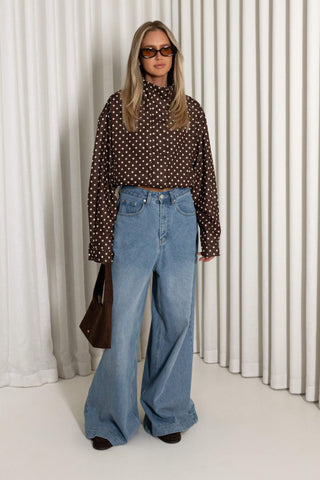A woman modeling a brown long-sleeve blouse with a white polka-dot pattern, paired with high-waisted, wide-leg light blue jeans, sunglasses, and a dark brown handbag, set against a background of white vertical drapes.