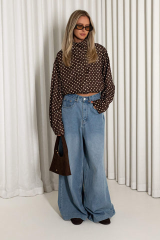 A woman modeling a brown long-sleeve crop top with a white polka dot pattern, paired with high-waisted wide-leg blue jeans, sunglasses, and a dark brown handbag, standing in front of a white curtain backdrop.