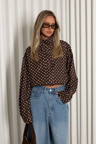 A woman modeling a brown long-sleeve crop top with a white polka dot pattern, paired with light blue jeans and dark sunglasses, standing in front of a white curtain background.