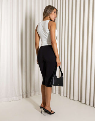 Woman in a white sleeveless top and black capri pants holding a black handbag against a white curtain background. Dustin white sleeveless V-neck top with gold hardware
