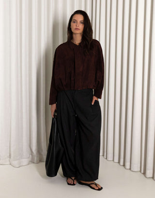 Person wearing a dark brown bomber and black balloon pants standing against a white curtain. black balloon leg pants