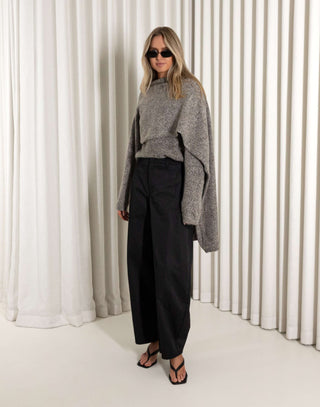 Woman wearing a gray sweater and black pants standing against a white curtain.