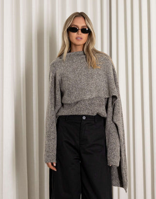 Woman wearing a gray sweater and black pants against a white curtain background