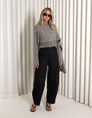 Woman wearing a gray knit and black pants against a white curtain background