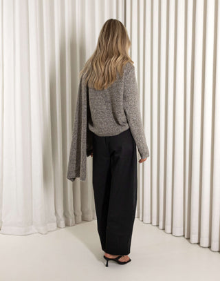 Person wearing a gray knit and black pants standing in front of white curtains.