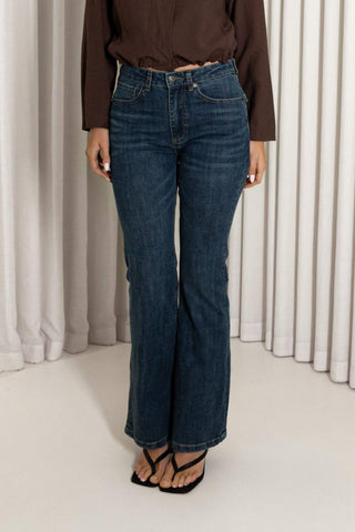 A person wearing dark blue high-rise flare jeans, a brown long-sleeve top, and black flat sandals, standing in front of white draped curtains.