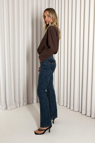 A woman modeling dark blue flare jeans with a brown long-sleeve top and black heels, standing in front of a white pleated curtain backdrop.