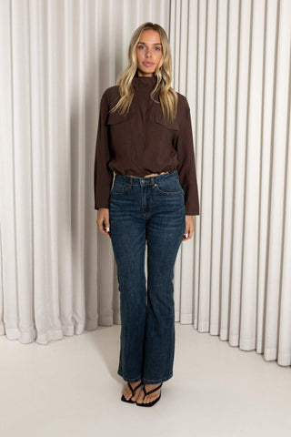 A woman stands in front of white pleated curtains wearing a dark brown long-sleeve mock-neck top, dark blue high-waisted flare jeans, and black strappy sandals.