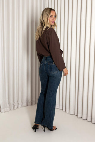 A woman wearing a brown long-sleeve top and dark blue flared jeans, standing in front of white pleated curtains and looking back over her shoulder.