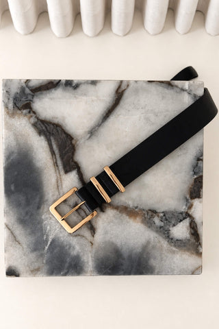 A black leather belt with gold-tone hardware, including a square buckle and two decorative rings, laid diagonally across a gray and white marble block.