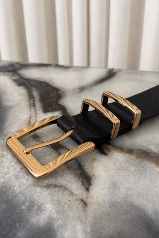 A close-up of a black leather belt with a textured gold buckle, resting on a gray and white marble surface with a soft-focus cream curtain background.