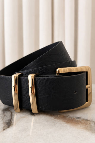 A black textured leather belt with polished gold-tone metal buckles, coiled on a light marble surface with a soft beige curtain background.