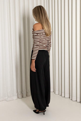 A woman stands with her back to the camera wearing a brown and white striped off-the-shoulder top and black wide-leg pants against a white pleated curtain backdrop.