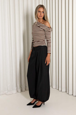 A woman modeling a brown and beige striped off-the-shoulder long-sleeve top paired with black wide-leg pants and black pointed-toe heels, standing in front of white vertical drapes.