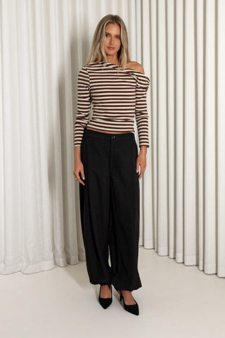 A woman modeling a brown and beige striped off-the-shoulder long-sleeve top paired with black wide-leg trousers and black pointed heels, standing in front of a white curtain backdrop.