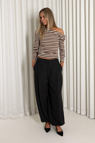 A woman modeling a brown and white striped off-the-shoulder long-sleeve crop top with black wide-leg pants and black pointed-toe heels, standing in front of a white curtain backdrop.
